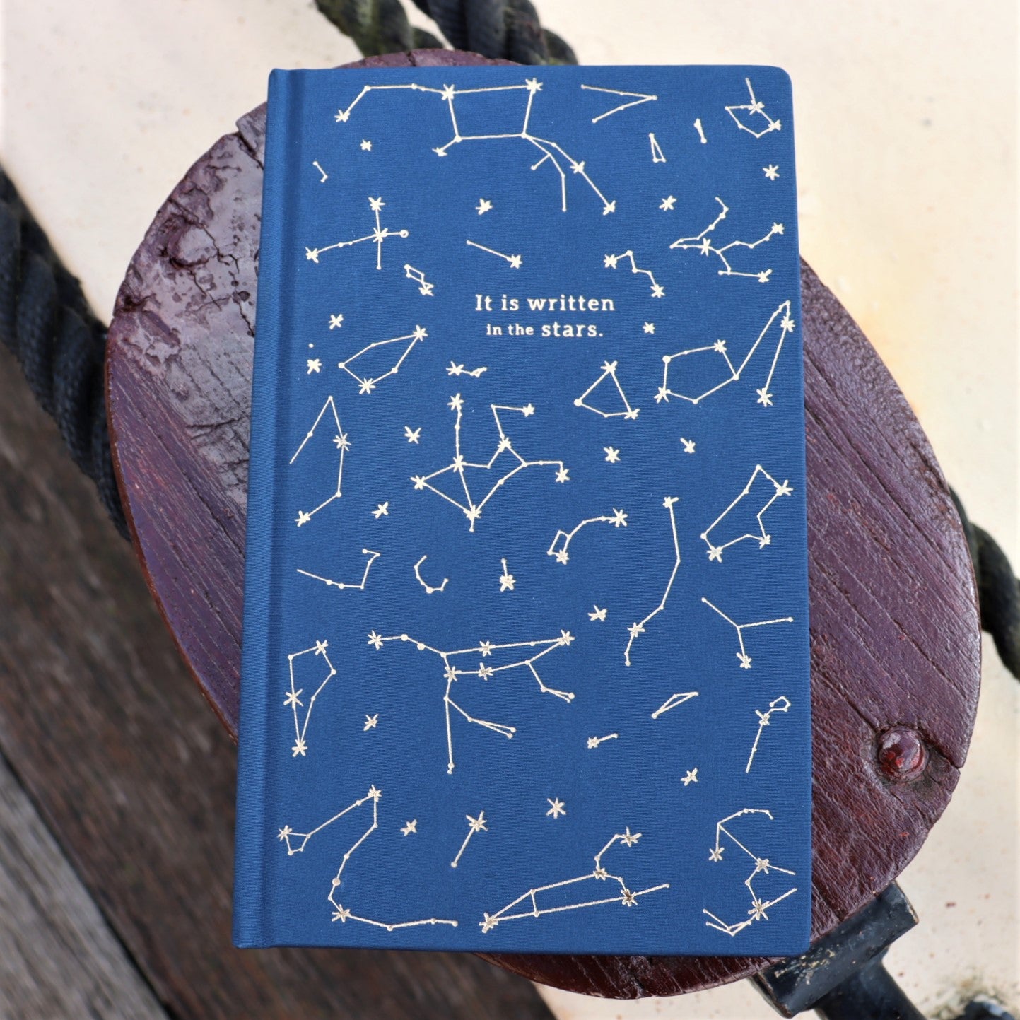 A blue clothbound journal with gold foil embossed constellations and the phrase 'It is written in the stars' on the cover, placed on a wooden surface.
