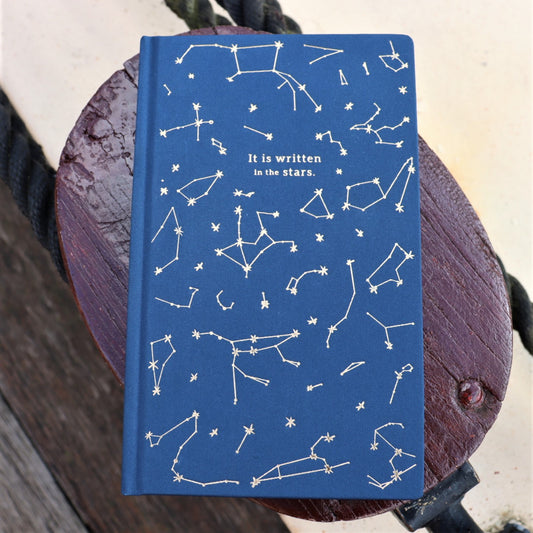 A blue clothbound journal with gold foil embossed constellations and the phrase 'It is written in the stars' on the cover, placed on a wooden surface.