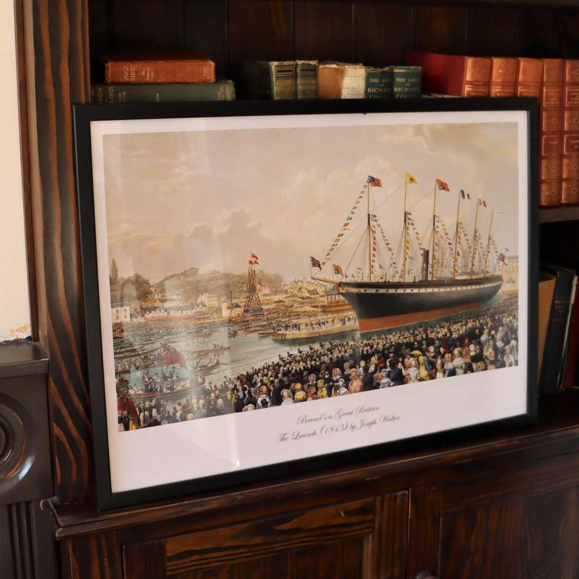 A framed replica print of Joseph Walter's 'The Launch' painting, depicting the launch of the SS Great Britain into Bristol's Floating Harbour, displayed on a wooden shelf.