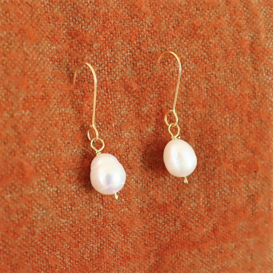 A pair of gold-plated hook earrings with large white pearl drops, displayed against a orange-brown background.