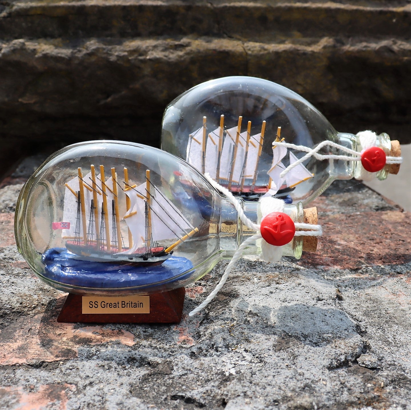 Two glass bottles with replica ships inside, one larger than the other, displayed on a stone surface with a 'SS Great Britain' label in front.