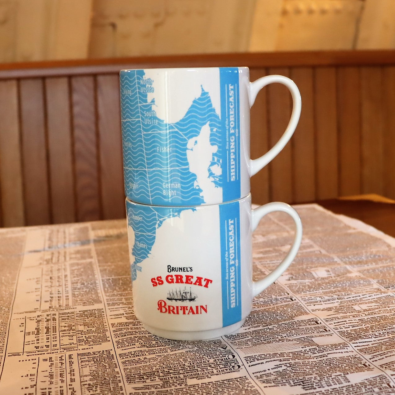 Two white stacking mugs with a blue shipping forecast design and the SS Great Britain logo, placed on a surface with a newspaper print.
