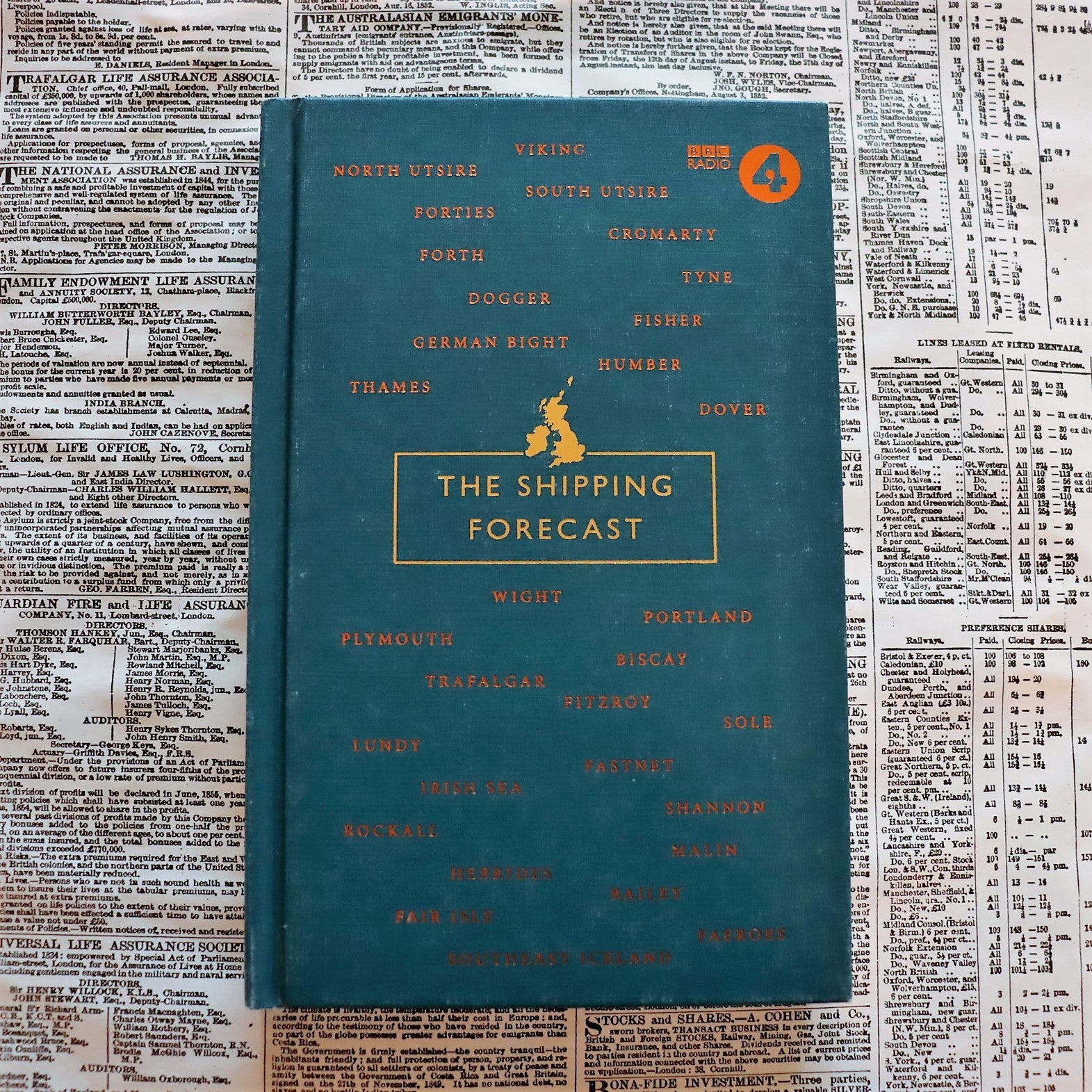 A hardcover book titled 'The Shipping Forecast' lying on a surface with text, featuring a blue cover with gold text and a map outline.