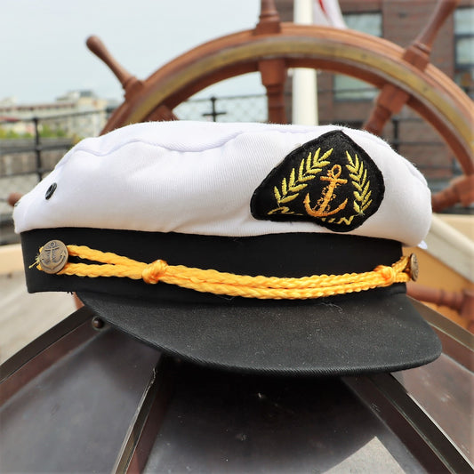 A captain's cap with a white front and black visor, featuring a gold anchor and laurel emblem, displayed on a wooden surface with a ship's wheel in the background.