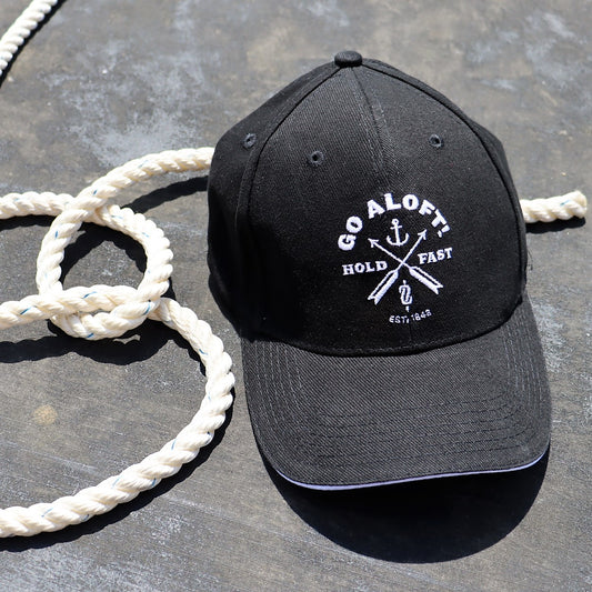 A black cap with the 'Go Aloft! Hold Fast' logo printed in white, placed on a surface with a coiled white rope beside it.