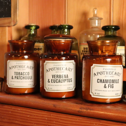 A set of three apothecary glass jar candles with different labels indicating Tobacco & Patchouli, Verbena & Eucalyptus, and Chamomile & Fig scents, arranged on a wooden surface.