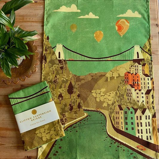 A green and yellow tea towel with an illustration of a suspension bridge and surrounding landscape, displayed on a wooden table with a plant in the background.