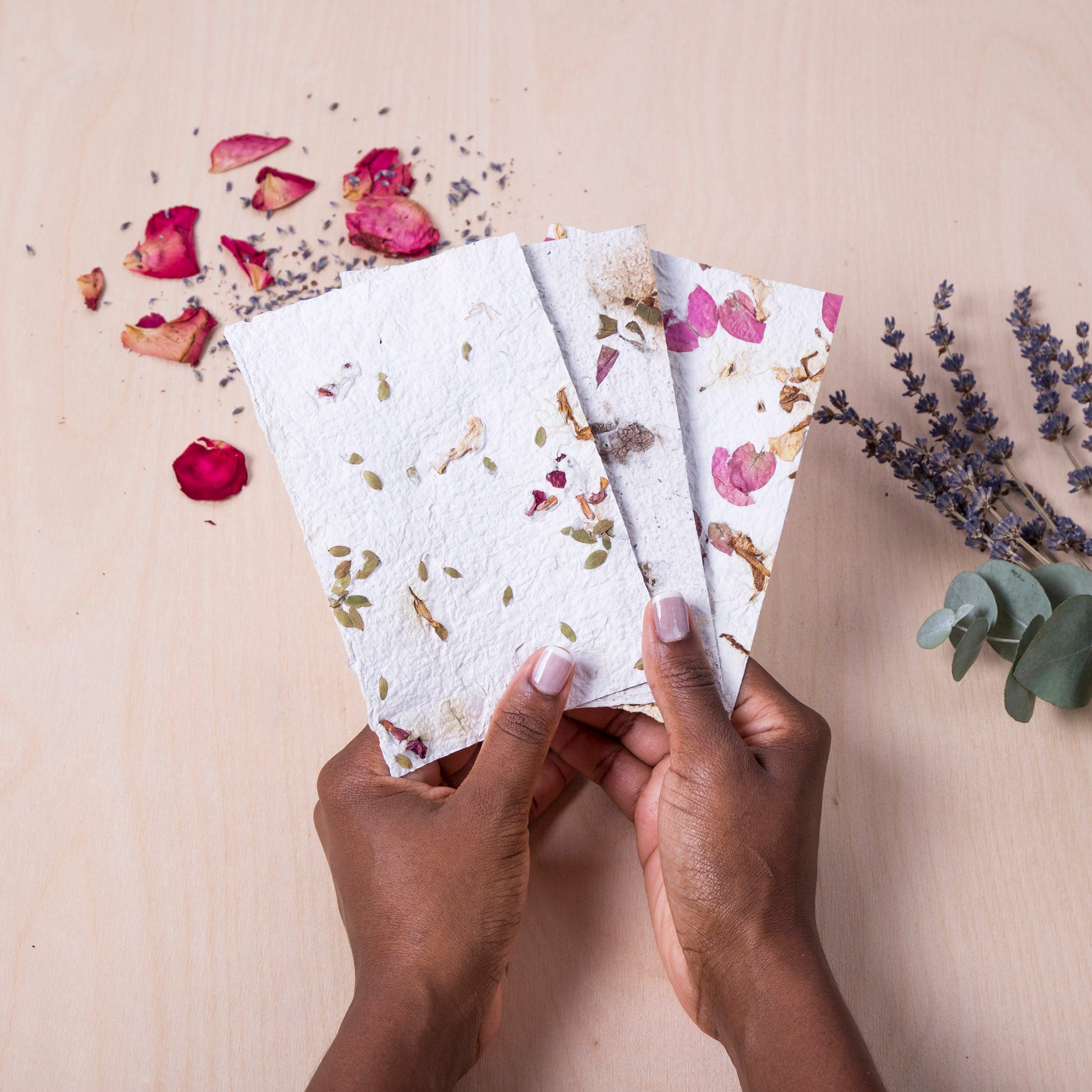 A person's hands holding handmade paper with pressed flowers, alongside scattered dried flowers.