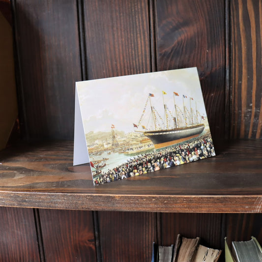 A greeting card with a historical watercolor illustration of the SS Great Britain's launch, placed on a wooden shelf.