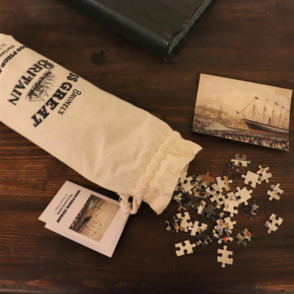 A 1000 piece jigsaw puzzle depicting the SS Great Britain, with pieces scattered on a surface and the puzzle packaged in a canvas bag with a printed illustration.